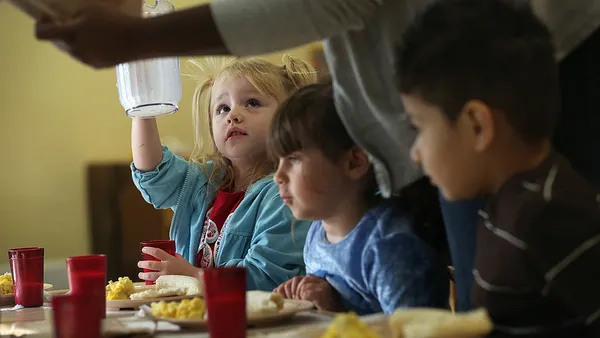 A young student is seated at a table holding an empty pitcher and looking up at a person who is standing. Only the arms and upper body of the person standing is shown. Other young students are seated at the table.