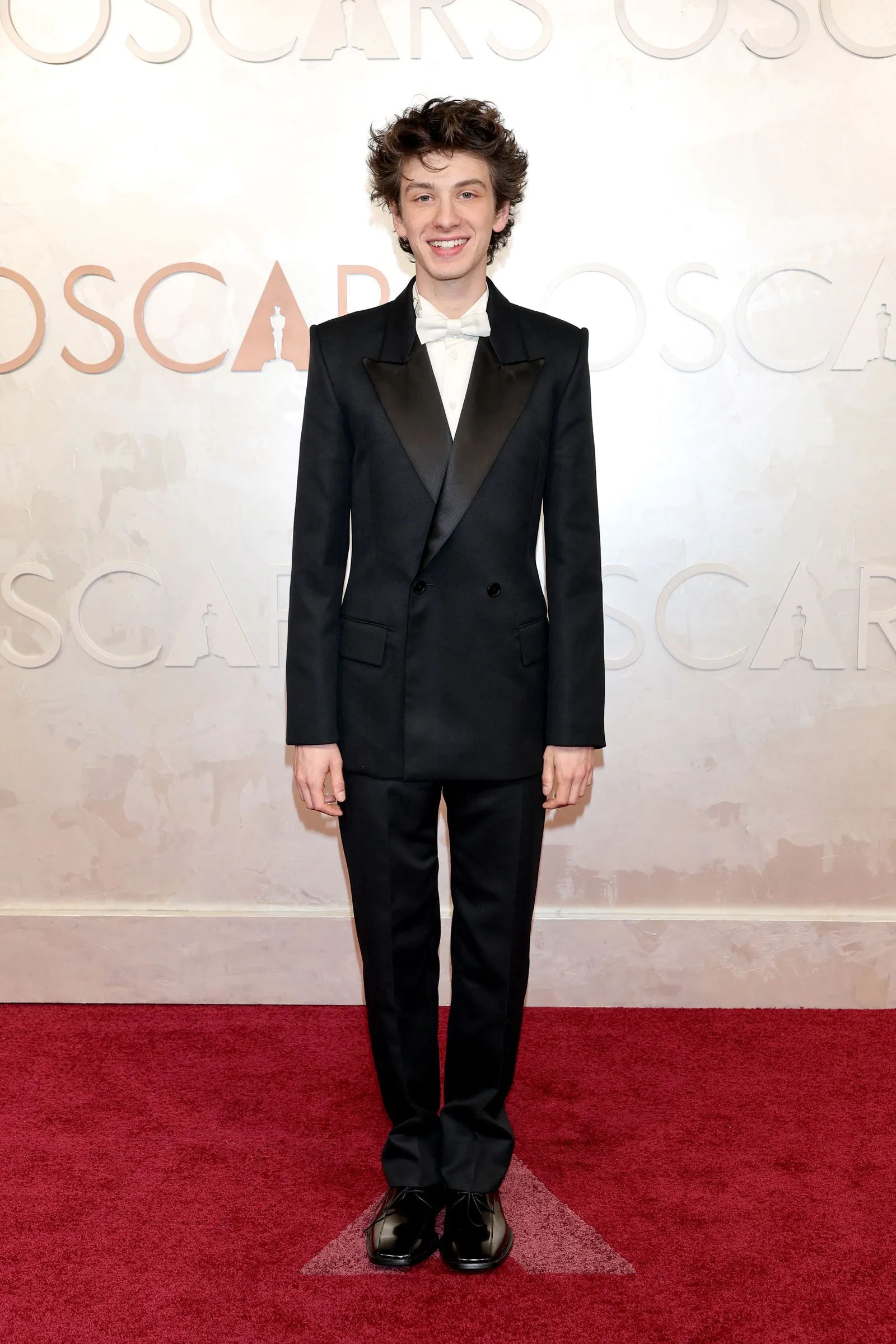 A man in a black tuxedo and white shirt and bow tie stands on the red carpet.