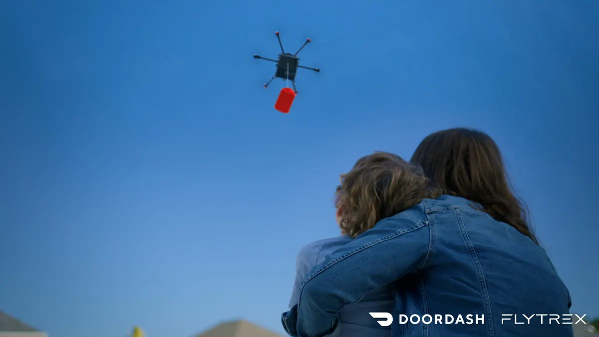 An image of an adult and child looking up at the sky where there is a drone carrying a red bag.