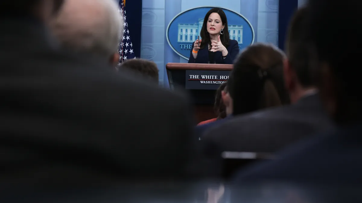 Woman speaks at a White House podium in the distance, seen through a blurred out crowd.