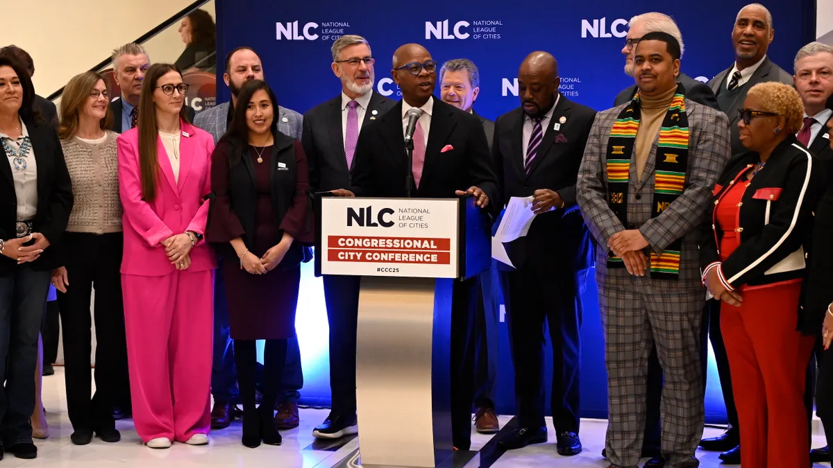 A man at a podium with a sign reading "National League of Cities Congressional City Conference" surrounded by other men and women.