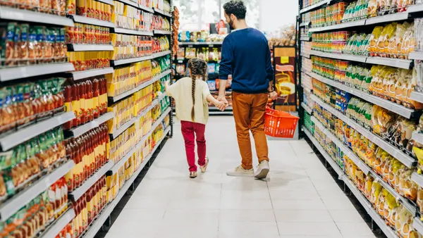 A child and parent are holding hands while walking down an aisle of a grocery store. The parent has a shopping basket.