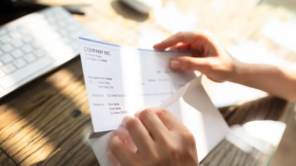 Close-up Of A Businessperson's Hand Opening Envelope With Paycheck Over Wooden Desk
