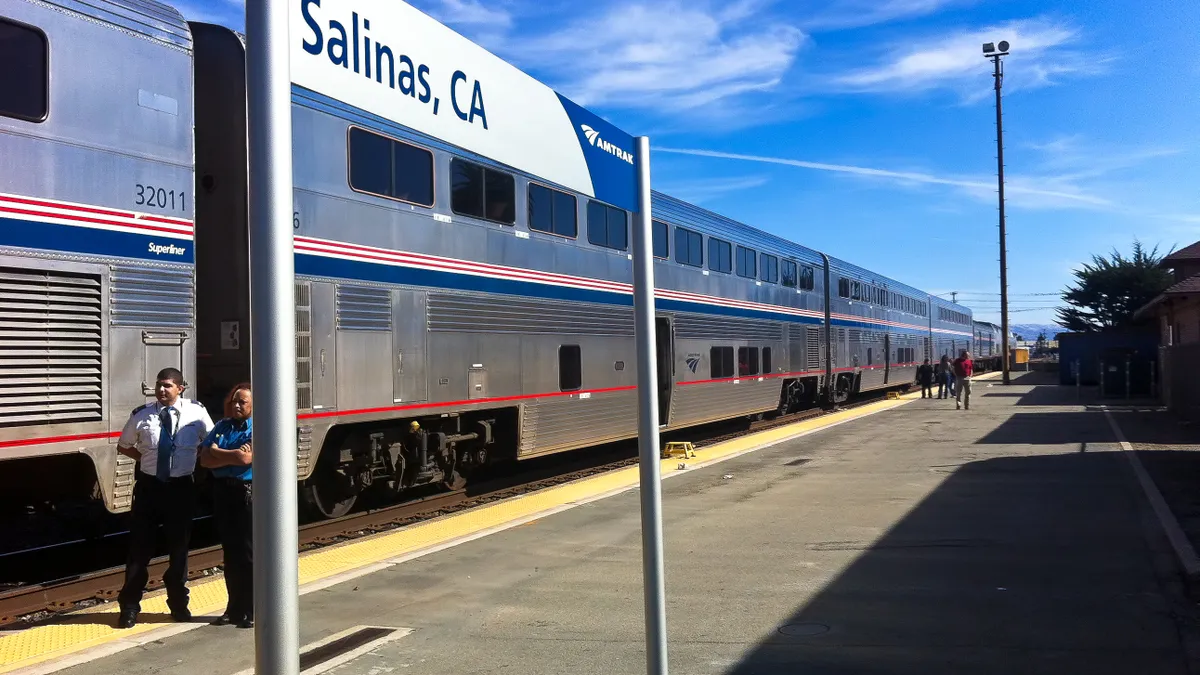 An Amtrak train is stopped at a station identified by a big sign in the foreground reading "Salinas" as two railroad workers stand on the platform.