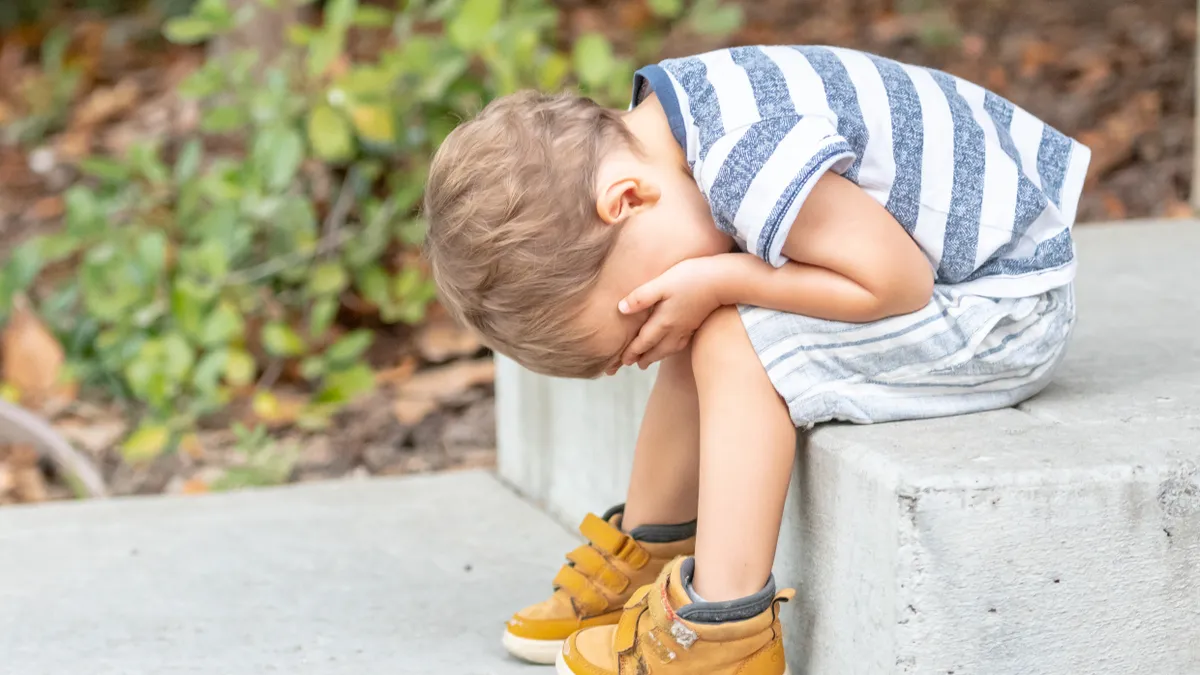 A young child sits on a concrete step with head in lap.