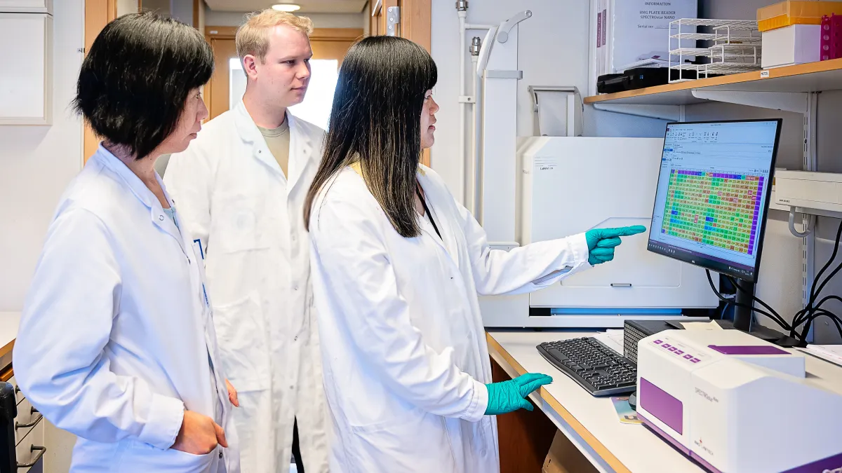 Landscape photo of three researchers, one of whom is pointing at a computer screen, looking at data in a laboratory setting.