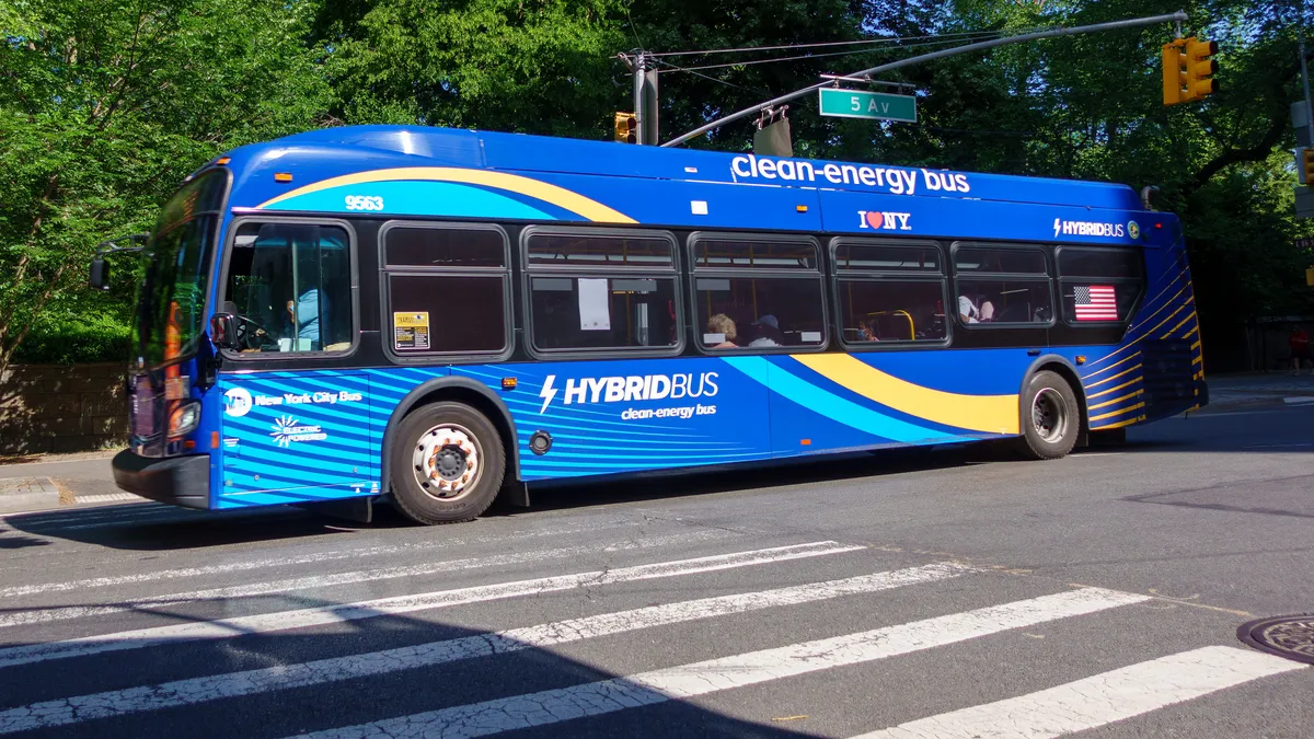 A blue New York City transit bus marked "clean-energy bus" travels along the curb lane under a sign for Fifth Avenue, with trees in the background.