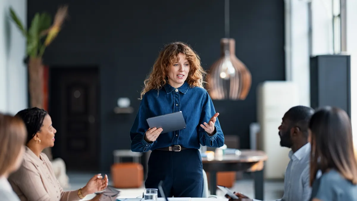 A woman stands at a podium, presenting to a group of attentive colleagues seated in a conference room.