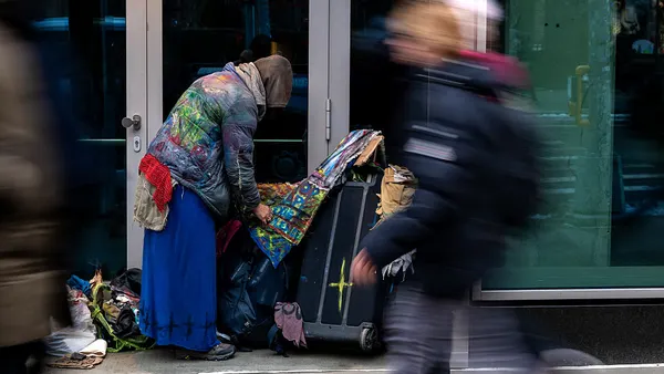 A person standing and facing away from the camera is wearing a coat with a hood while touching a cloth draped over an upright suitcase.