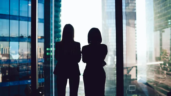 Back view of female colleagues in formal wear standing near window looking at modern exterior of skyscrapers in business center.