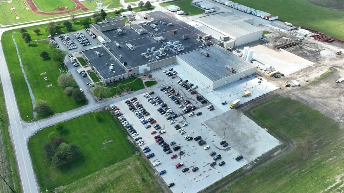 The drone view of a building next to a parking lot with cars, surrounded by green grass.