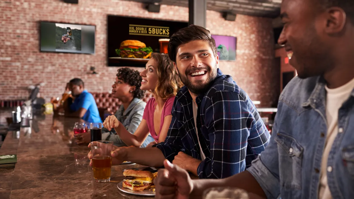 Five people sitting down at a bar talking and laughing. One man has a drink and a burger.