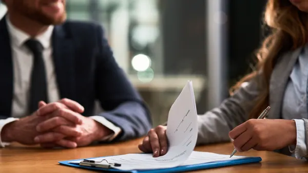 Two business people sitting at a desk, one is looking at a clipboard of papers and has a pen in hand.