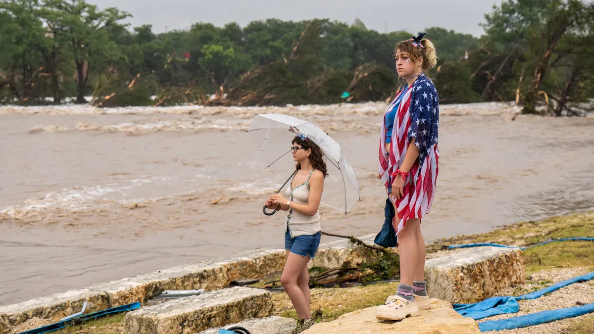 A person with an umbrella and a person wearing a stars-and-stripes shirt stand on rocks looking at muddy river water