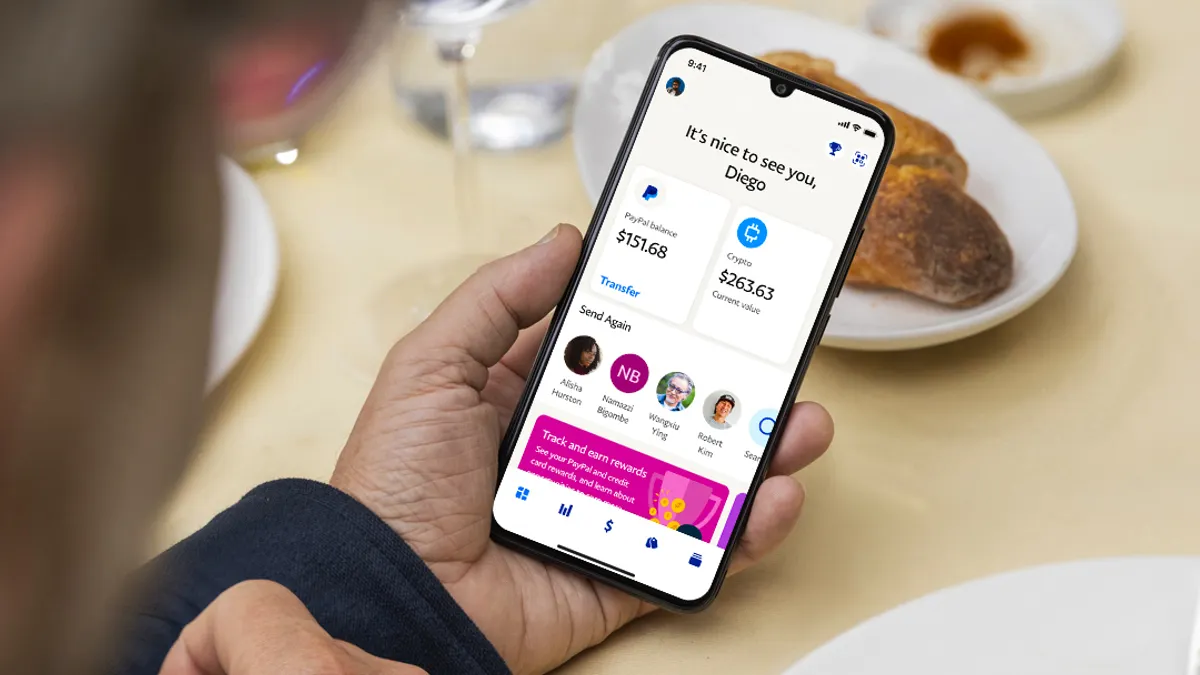 A hand holds a phone on a kitchen table. The screen reads, "It's nice to see you, Diego! PayPal balance: $151.68, Crypto: $263.63. Send again."