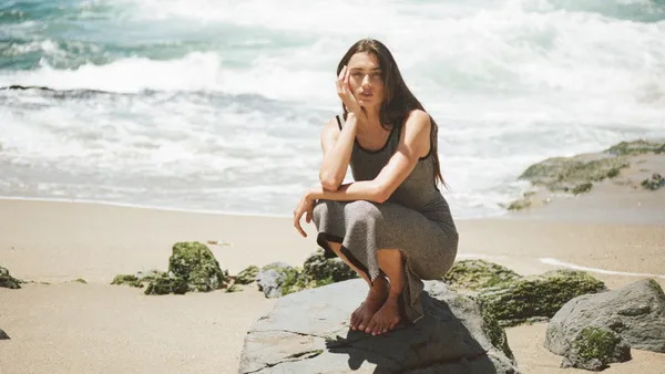 A person with long brown hair in a gray tank dress squats on a rock, on a sandy shoreline, during the day. Her head rests on one hand.