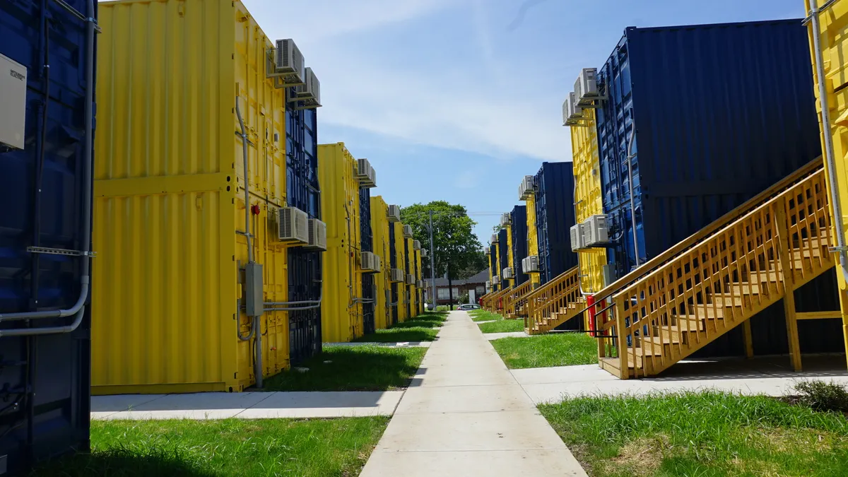 A white sidewalk with grass lawns and stacks of blue and yellow shipping container apartments on either side.