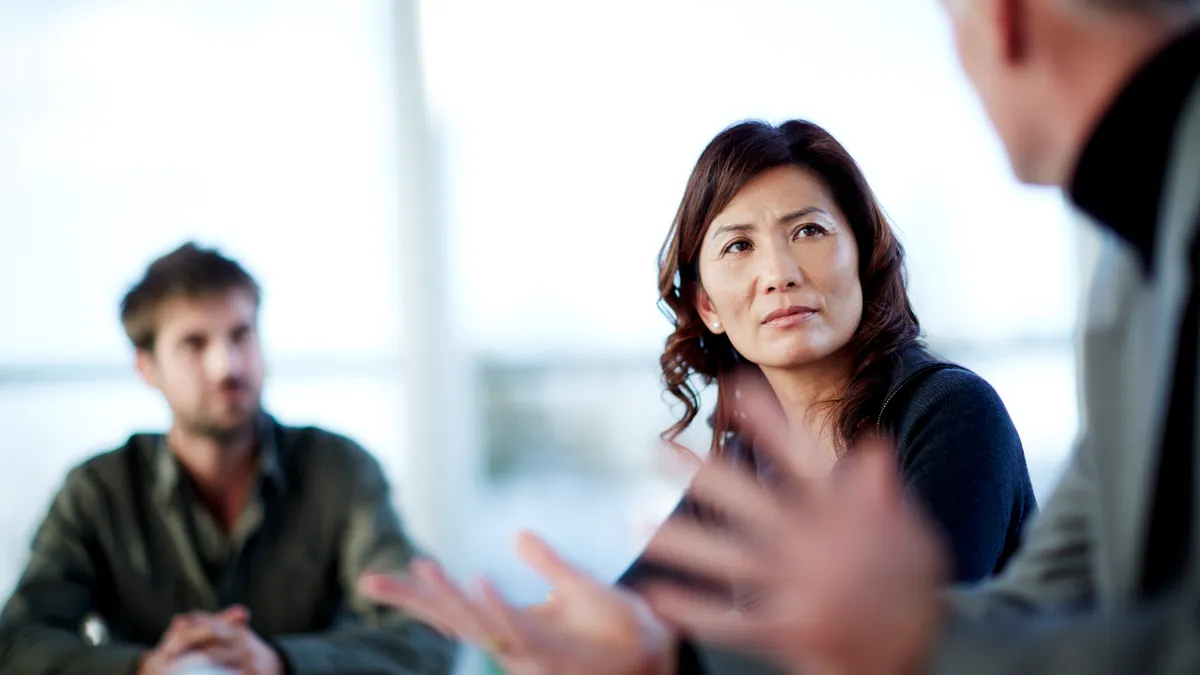 Pictured is a team meeting, where two White men are blurred and the Asian woman's face is in focus. She is in distress.