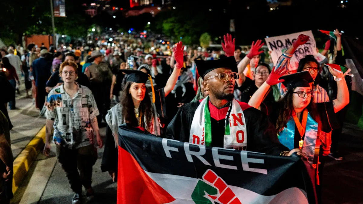 A crowd of people in graduation regalia, some with their hands painted red, march down a city street at night. The person in the center foreground holds a flag that reads "Free Palestine."