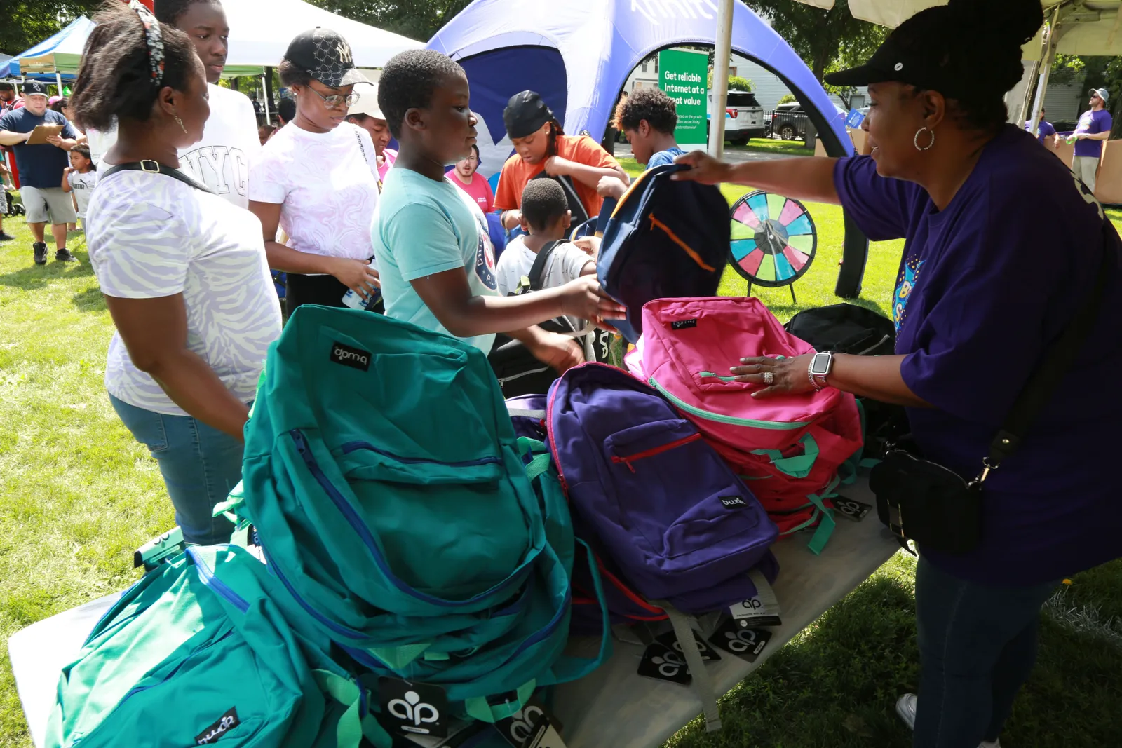 An adult standing under an outdoor tent and behind a table hands backpacks to several students