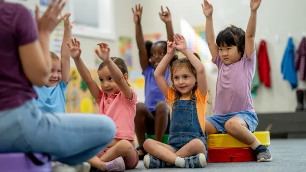 A group of young students is sitting on the floor in a classroom with their arms stretched above their heads. They are facing an adult who is sitting on the floor but it out of the camera shot and only their leg is visiable.