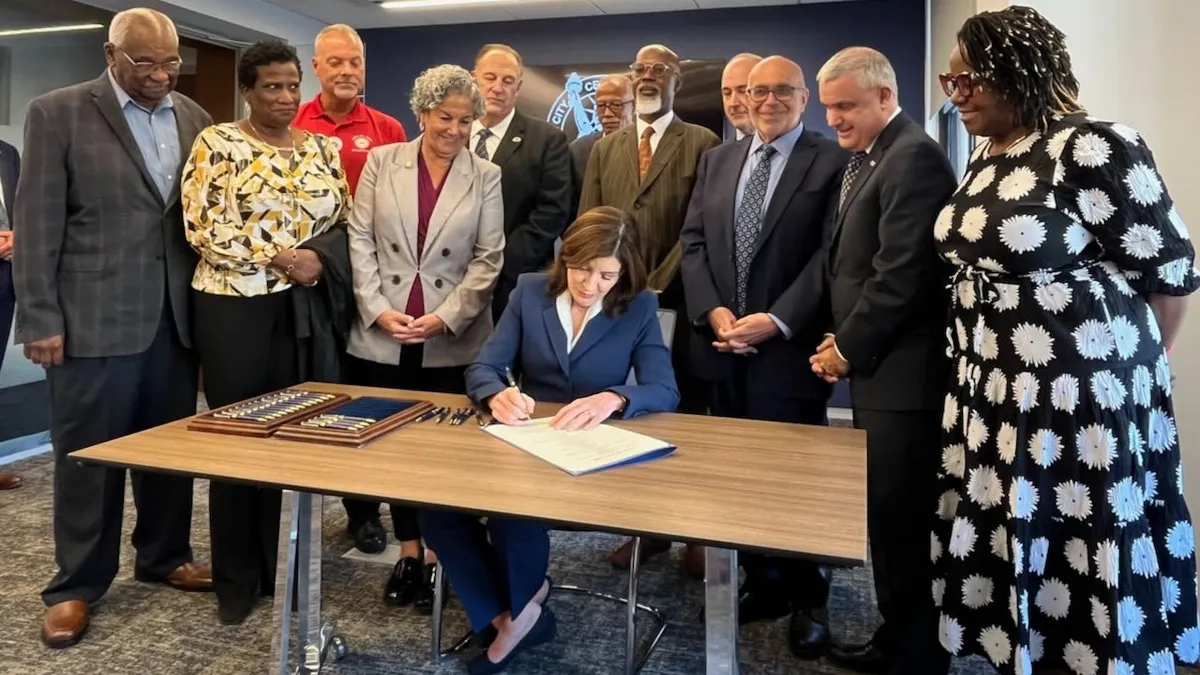 A diverse group of people stand around a table where a person in a blue pantsuit signs papers.