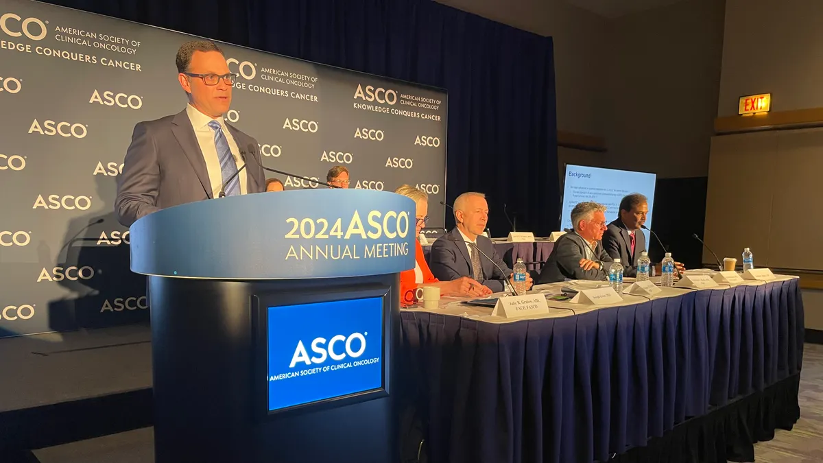 A person speaks at a podium next to four people seated at a lower table.