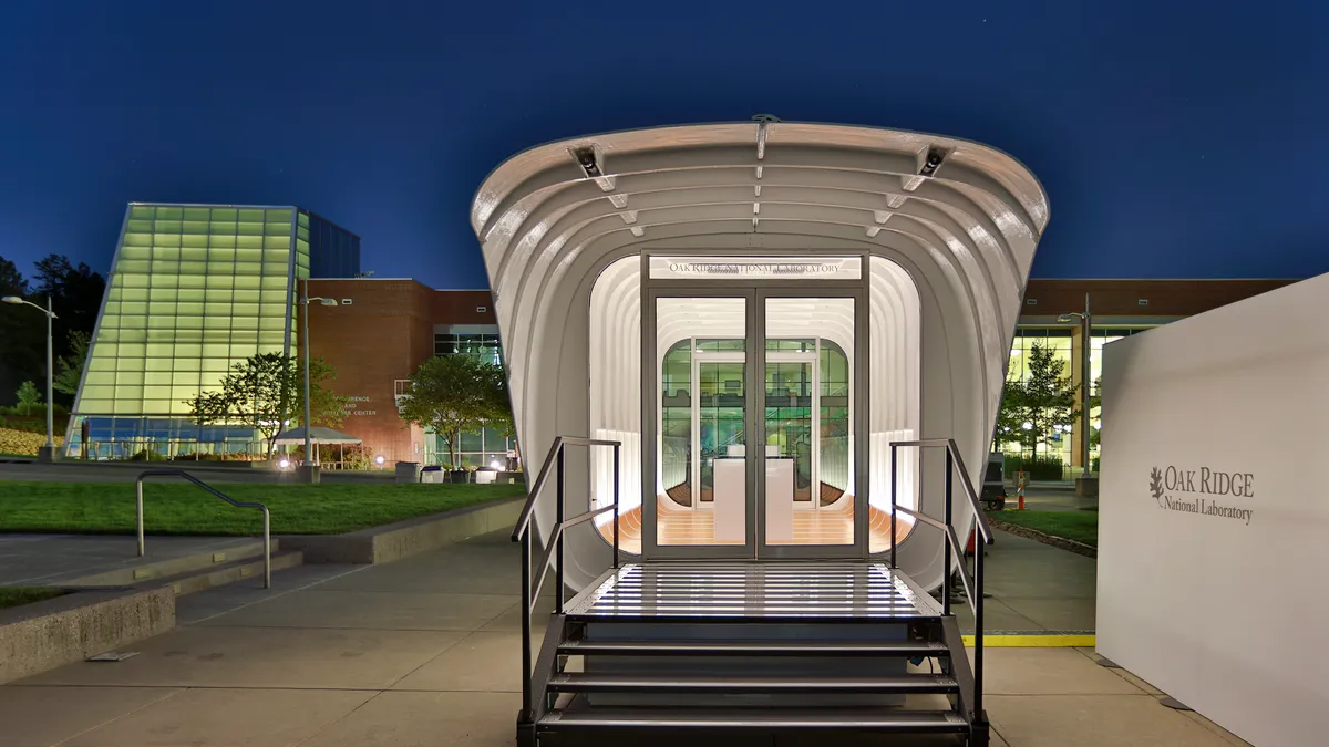 A white tunnel lit with glass doors and a white wall with Oak Ridge National Laboratory text on the right, and a brick building behind in the dusk.