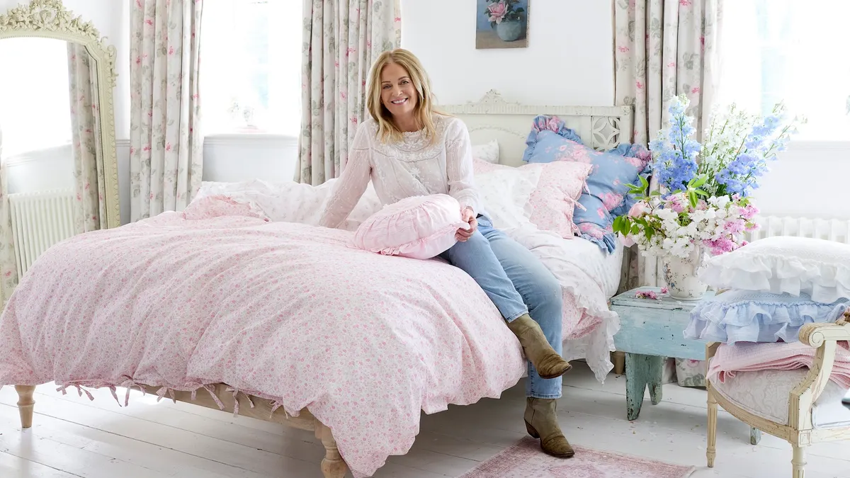 A person with long blond hair wearing jeans and ankle boots sits on a bed with a pink spread.