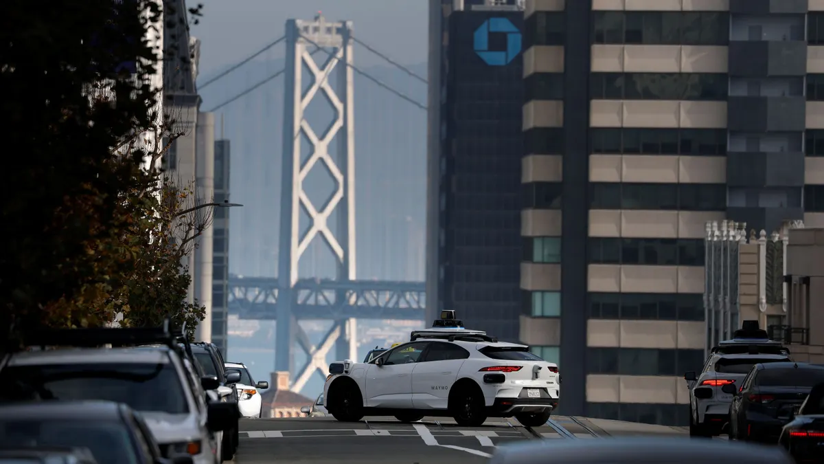 A Waymo robotaxi drives across an intersection in San Francisco, with the Bay Bridge and a building with a Chase logo towering in the background.