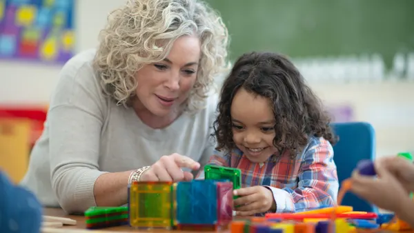 An adult and young student are sitting at a table. They are building a structure out of colorful magnet tiles.