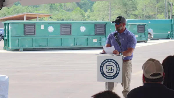 A man speaks at a poduim, standing in front of a row of containers meant for drop-off recycling