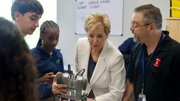 Two students and two adults stand near a table in a classroom and look at robotics with wheels