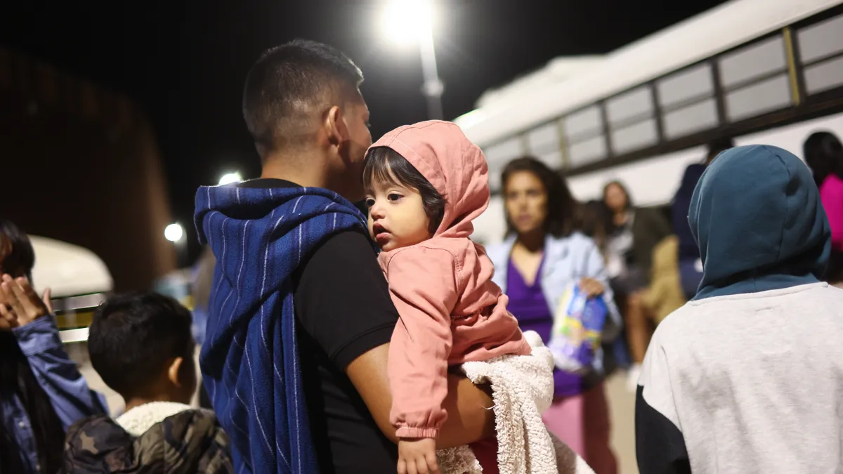 An adult, with back toward camera, holds a young child who is looking off to the side. They are outside and it's dark.