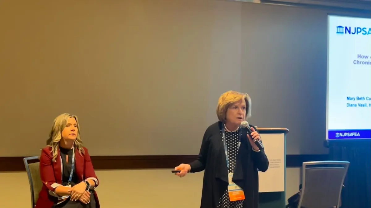 A presenter stands and talks in front of a principal who is sitting down during a conference session.