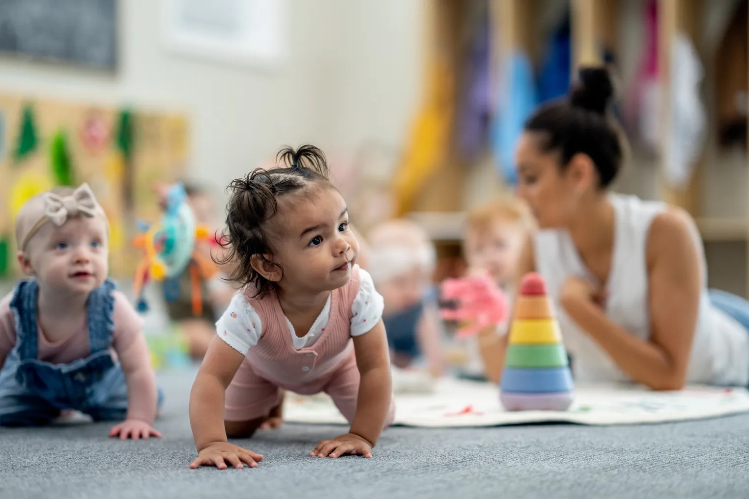 Two babies are in a crawl position on the floor looking to the side and ahead. In the background, an adult is on the floor with other children.