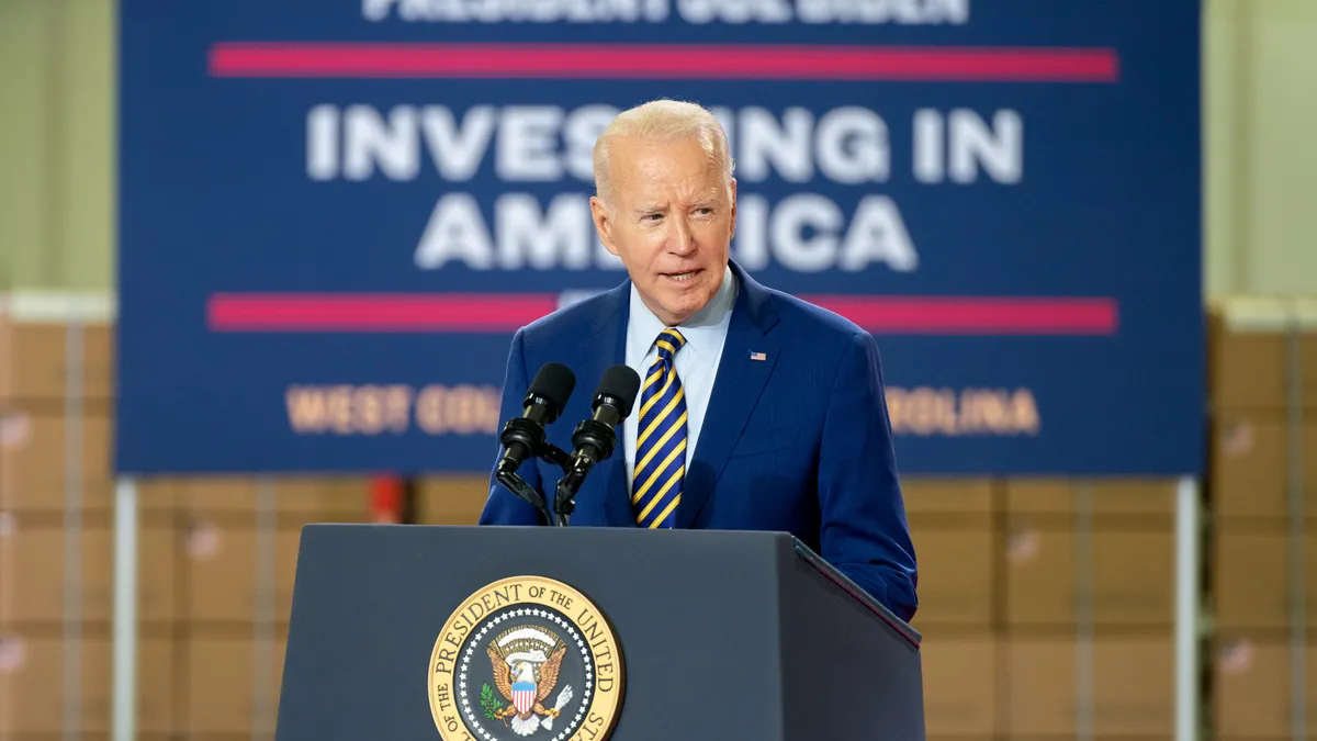 President Joe Biden in a blue blazer, white shirt and blue and yellow striped tie, standing behind a podium with a blue "Investing in America" sign blurred in the background.