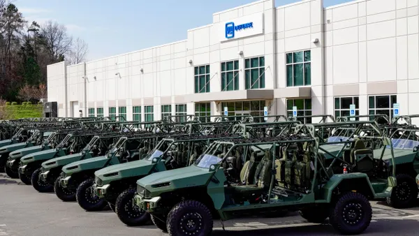 Two rows of green infantry squad vehicles parked in front of a white building with the blue GM Defense logo on it.