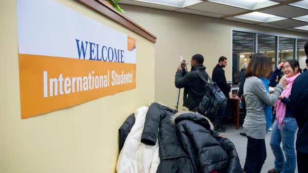 In a large room, a sign in the foreground reads "Welcome International Students!". Multiple groups of students chat amongst themselves in the background.