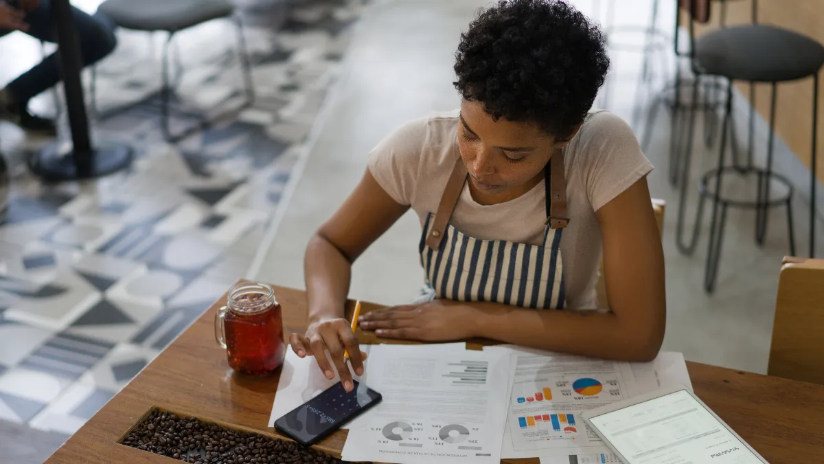 A woman sitting at a table using her phone as a calculator. On the table there are financial charts.