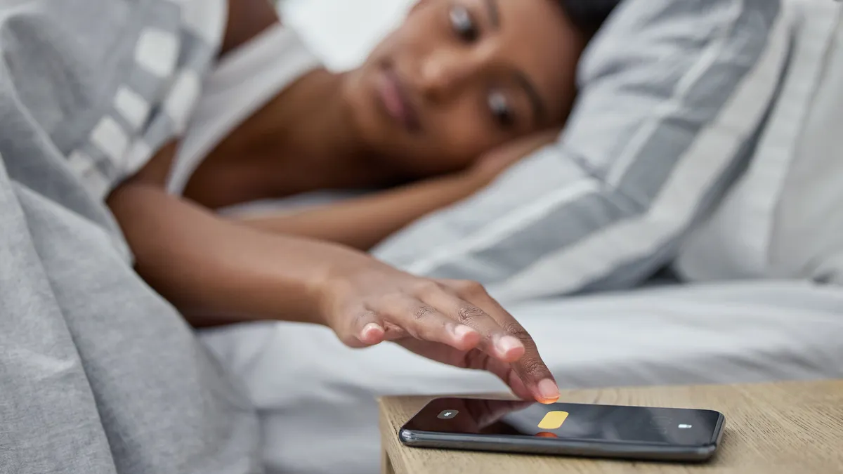 Person lays in bed while touching a smartphone on nightstand.