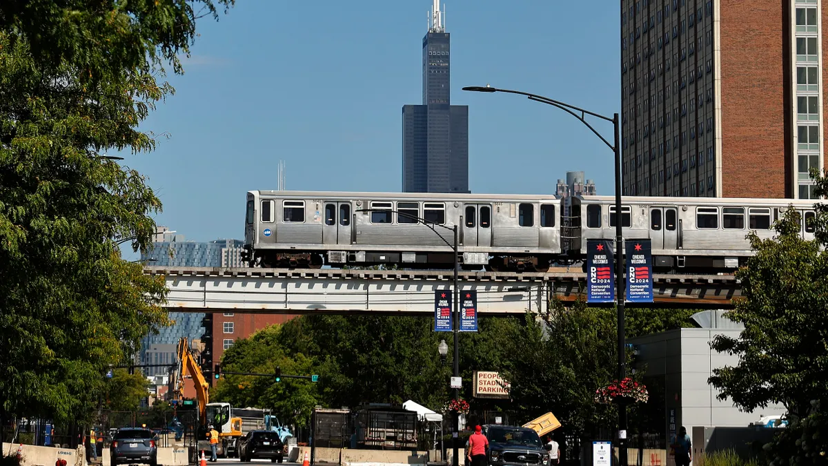 A silver elevated train over a city street with a tall building in the background.