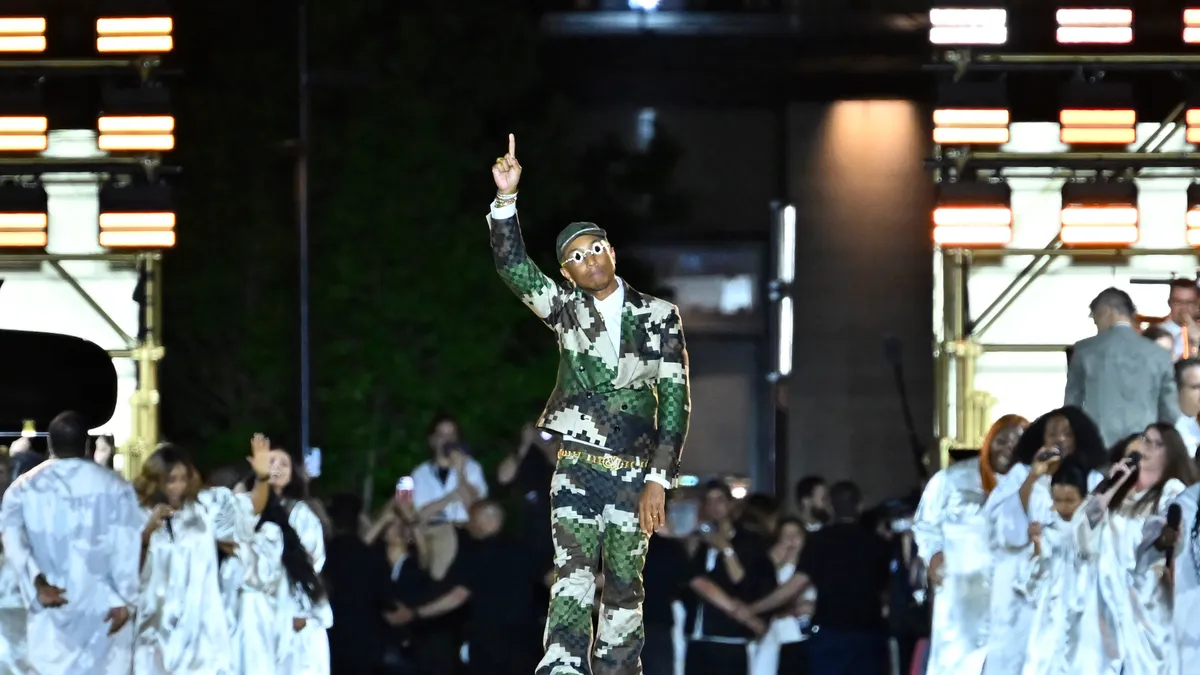 Pharrell Williams wears a green pixelated camo suit and points at the sky on the runway of his debut collection for Louis Vuitton menswear.