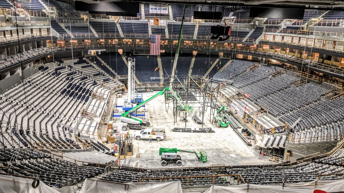 The inside of a stadium is being built. Workers are putting together the floor for a basketball court, and seats rise up around it. Equipment and material litters the floor.