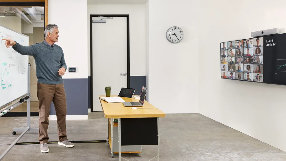 An adult stands in front of a white board while facing a wall with a television. On the television are squares of faces looking at the adult. In the middle of the room is a desk.