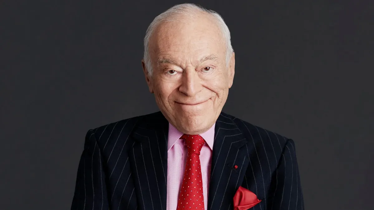 A man in a pin-stripe suit with a pink shirt and a red tie crosses his arms and smiles against a grey background.