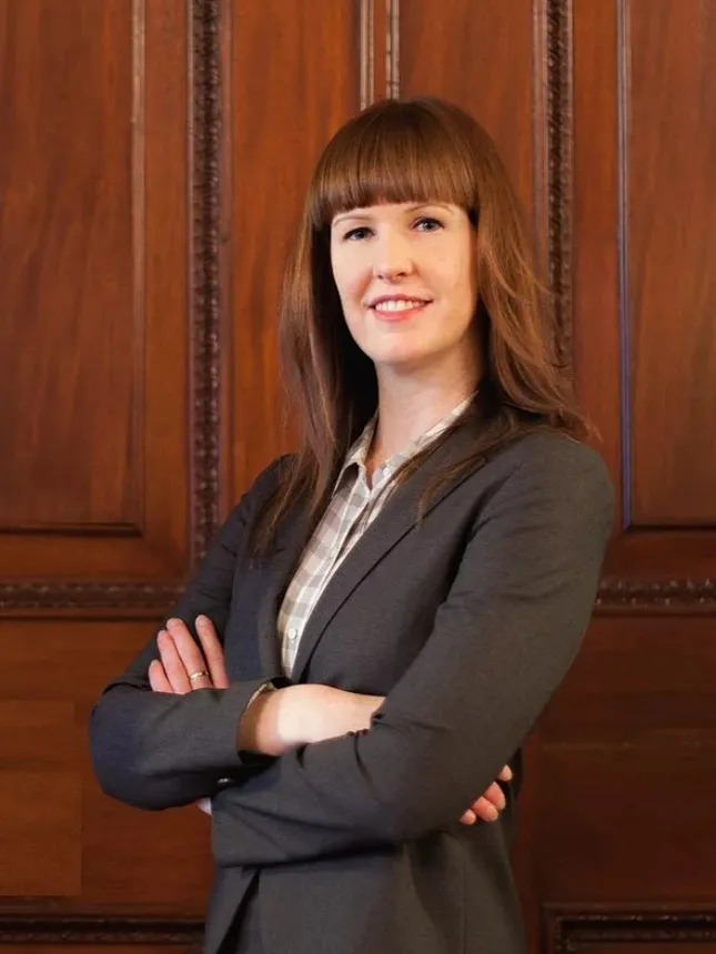 A woman wearing a suit stands in front of a wooden door with her arms crossed.