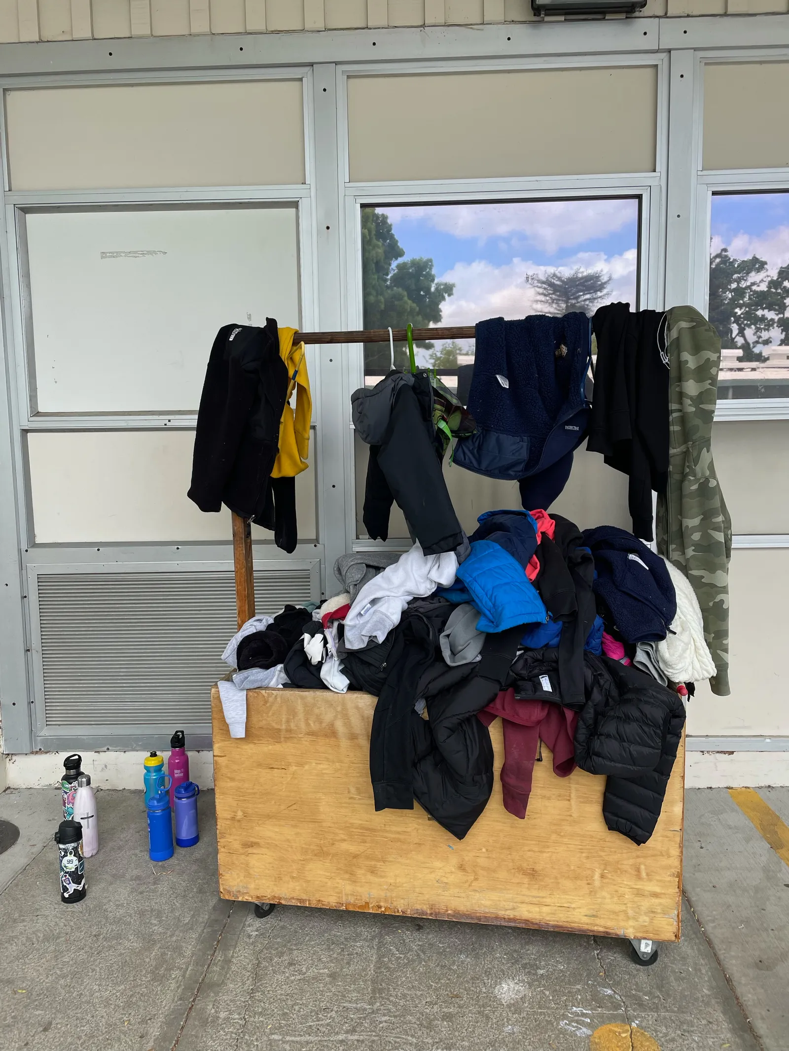 A clothing rack sits outside displaying clothes. A wooden cart also is filled with clothes. Several water bottles stand on the ground near the wooden cart.