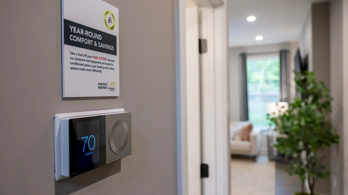 The hallway of a home with white walls, with a thermostat that reads "70" on the wall. Above it is a panel that reads "Year-Round Comfort & Savings."