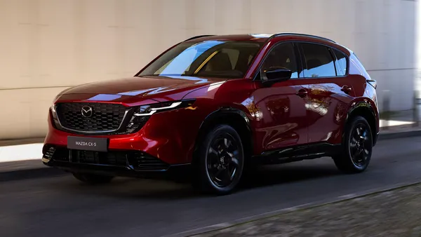 A red 2026 Mazda CX-5 crossover driving on a road with a concrete wall in the background.